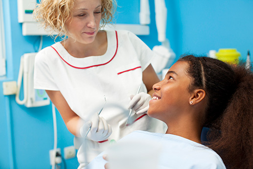 Young girl sitting in dentist chair with female tech holding dental tools.